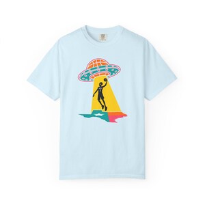 May include: Light blue t-shirt featuring a graphic of a basketball player being beamed up by a UFO over a Texas-shaped design. The UFO is orange and blue, and the player is black. The Texas design is green, pink, and blue.