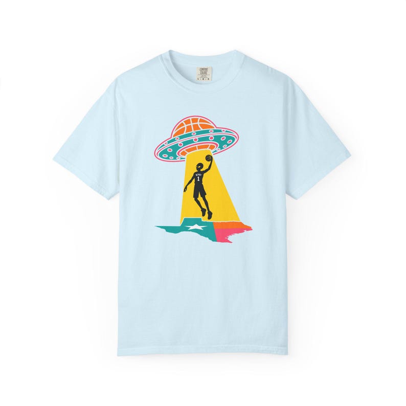 May include: Light blue t-shirt featuring a graphic of a basketball player being beamed up by a UFO over a Texas-shaped design. The UFO is orange and blue, and the player is black. The Texas design is green, pink, and blue.