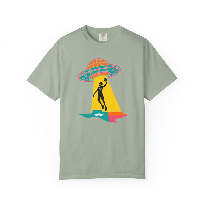 May include: Sage green t-shirt featuring a graphic of a basketball player being beamed up by a UFO. The UFO is orange and teal, with a pink outline. The player is black, and the beam is yellow. The Texas state outline is below.