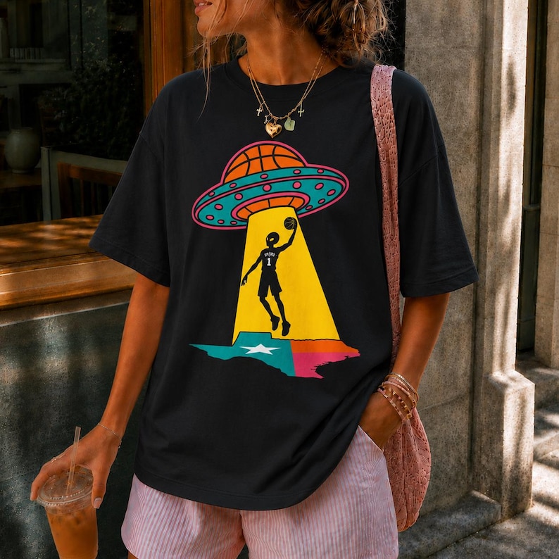 May include: Black t-shirt featuring a basketball-themed graphic. The design includes a basketball-shaped UFO, a basketball player, and a Texas-shaped outline. The graphic is in bright colors, with a yellow beam of light shining down on the player.