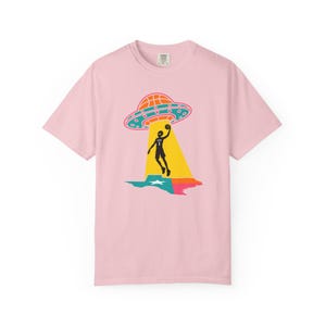 May include: A light pink t-shirt featuring a graphic of a basketball player being beamed up by a UFO over a colorful Texas outline. The UFO is orange and blue, and the player is black. The shirt has a crew neck and short sleeves.