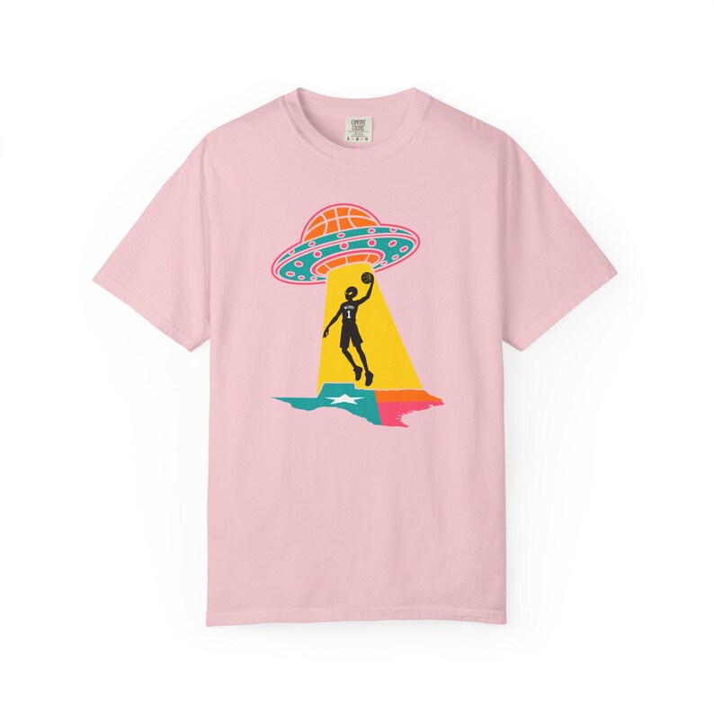 May include: A light pink t-shirt featuring a graphic of a basketball player being beamed up by a UFO over a colorful Texas outline. The UFO is orange and blue, and the player is black. The shirt has a crew neck and short sleeves.