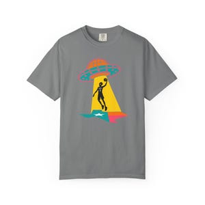 May include: Gray t-shirt featuring a graphic of a basketball player being beamed up by a UFO over a Texas-shaped design. The UFO is orange and pink, the player is black, and the Texas design is blue, green, and pink.