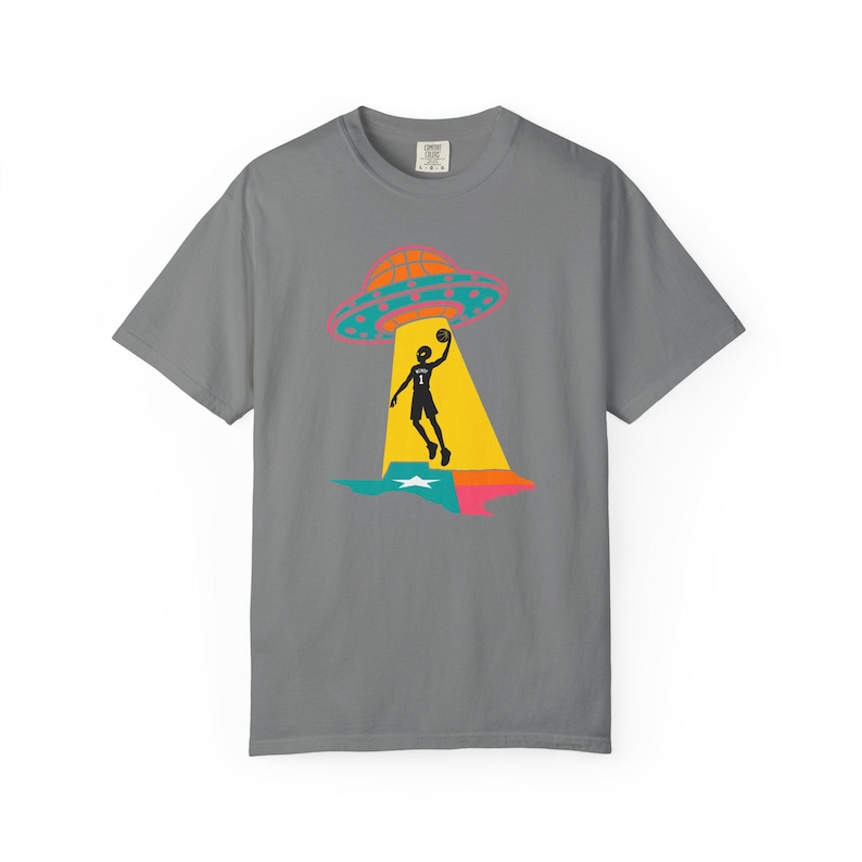 May include: Gray t-shirt featuring a graphic of a basketball player being beamed up by a UFO over a Texas-shaped design. The UFO is orange and pink, the player is black, and the Texas design is blue, green, and pink.