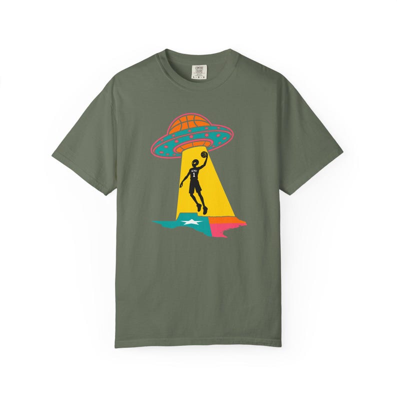 May include: Olive green t-shirt featuring a basketball-themed graphic. The design includes a basketball-shaped UFO, a basketball player, and a colorful outline of the state of Texas. The graphic is in black, orange, pink, yellow, and blue.