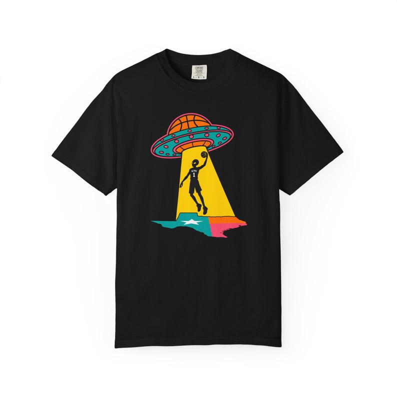 May include: Black t-shirt featuring a colorful graphic of a basketball player being beamed up by a UFO. The UFO has a basketball design and is above a Texas-shaped design with a star. The player is silhouetted in black.