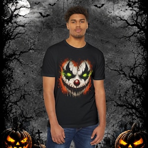 May include: A black short-sleeve t-shirt featuring a graphic print of a sinister clown face with glowing green eyes, a red nose, and a wide, jagged smile surrounded by orange and black splatters on the front center of the garment.