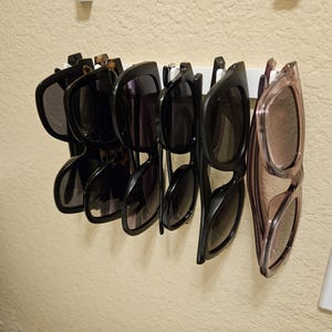 Puede incluir: Una colección de gafas de sol colgadas en un estante de pared. Las gafas de sol son una mezcla de monturas negras, marrones y transparentes con lentes oscuros.