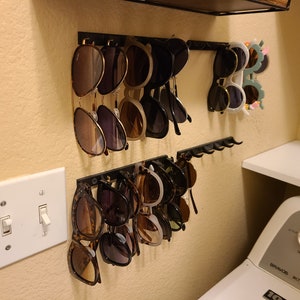 Peut inclure: Un organisateur de lunettes de soleil mural avec plusieurs paires de lunettes de soleil accrochées à des crochets. L'organisateur est en métal noir et a un design simple. Les lunettes de soleil sont un mélange de styles et de couleurs, y compris le brun, le noir et le blanc.