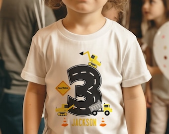 May include: A white t-shirt featuring a black number 3 surrounded by a black road with yellow lines. There are yellow construction vehicles on the road, including a dumper truck, an excavator, and a traffic cone. The text "CAUTION" is on a yellow diamond-shaped sign. The text "JACKSON" is below the number 3.