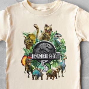May include: Cream-colored short-sleeved t-shirt featuring a colorful dinosaur graphic. The design includes various dinosaurs, the name "ROBERT", and the number "5" in a stylized font, set against a green foliage backdrop.