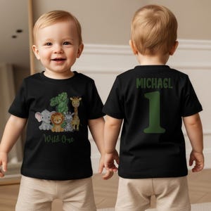 Pu&ograve; includere: T-shirt nero per bambini piccoli con una grafica di animali della giungla e la scritta "Wild One". La parte posteriore della maglietta ha il nome "MICHAEL" e il numero "1" in verde. Il bambino indossa pantaloncini color cachi.