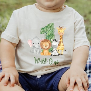 Pu&ograve; includere: T-shirt color crema per bambini piccoli con un design di animali della giungla. Il design include un elefante, un leone, una zebra e una giraffa, con il numero "1" e le parole "Wild One" in verde. La maglietta &egrave; in materiale morbido.