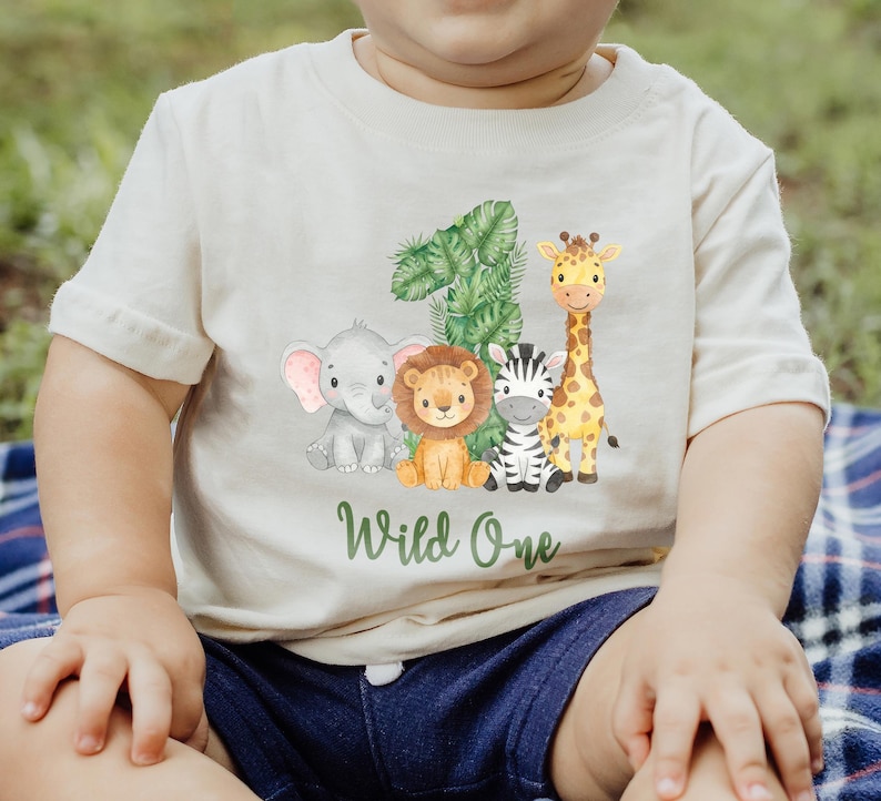 Pu&ograve; includere: T-shirt color crema per bambini piccoli con un design di animali della giungla. Il design include un elefante, un leone, una zebra e una giraffa, con il numero "1" e le parole "Wild One" in verde. La maglietta &egrave; in materiale morbido.
