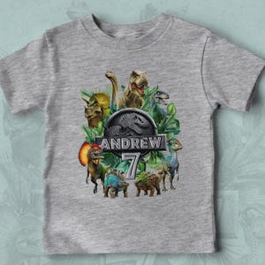 Peut inclure: Un t-shirt gris avec un motif color&eacute; de dinosaures, dont un T-Rex et un Tric&eacute;ratops, entour&eacute;s de feuillage vert. Le design comprend le nom "ANDREW" et le chiffre "7" dans une police stylis&eacute;e.