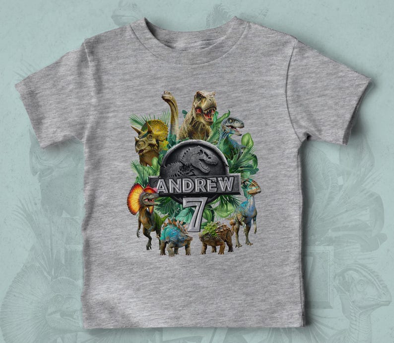 Peut inclure: Un t-shirt gris avec un motif color&eacute; de dinosaures, dont un T-Rex et un Tric&eacute;ratops, entour&eacute;s de feuillage vert. Le design comprend le nom "ANDREW" et le chiffre "7" dans une police stylis&eacute;e.