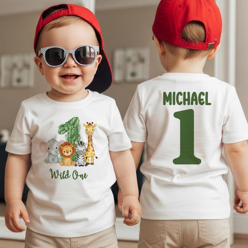 Pu&ograve; includere: T-shirt bianca con il numero 1 verde e il nome "Michael" sul retro. La parte anteriore presenta un design "Wild One" con un 1, elefante, leone, zebra e giraffa.