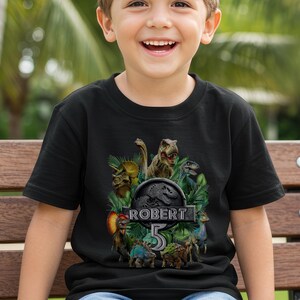 Peut inclure: T-shirt noir avec un motif de dinosaures color&eacute;s, le texte "ROBERT 5" et le logo Jurassic Park. Le design comprend diff&eacute;rents dinosaures. Ce t-shirt est probablement pour un anniversaire d'enfant.