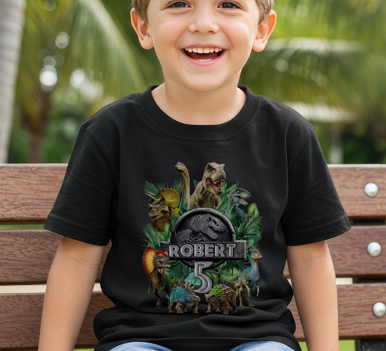 Peut inclure: T-shirt noir avec un motif de dinosaures color&eacute;s, le texte "ROBERT 5" et le logo Jurassic Park. Le design comprend diff&eacute;rents dinosaures. Ce t-shirt est probablement pour un anniversaire d'enfant.