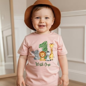 Pu&ograve; includere: Una maglietta rosa chiaro con il numero 1 e un motivo a foglie verdi, pi&ugrave; animali dei cartoni animati: un elefante, un leone, una zebra e una giraffa. Le parole "Wild One" sono stampate sotto. Il bambino indossa un cappello marrone.