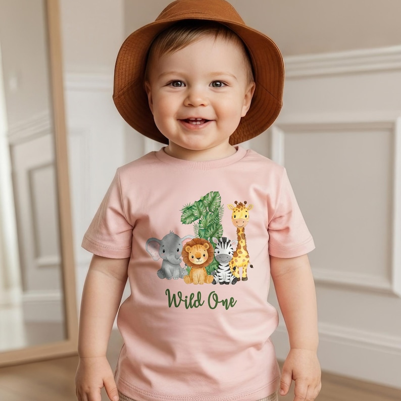 Pu&ograve; includere: Una maglietta rosa chiaro con il numero 1 e un motivo a foglie verdi, pi&ugrave; animali dei cartoni animati: un elefante, un leone, una zebra e una giraffa. Le parole "Wild One" sono stampate sotto. Il bambino indossa un cappello marrone.