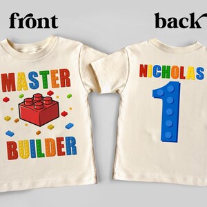 May include: Cream-colored toddler t-shirt with a colorful graphic. The front says "MASTER BUILDER" with a red block design. The back has the name "NICHOLAS" and a blue number "1".