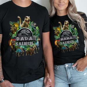 Peut inclure: T-shirts noirs avec des graphiques de dinosaures. L'un des t-shirts dit "DADA SAURUS" et l'autre "MAMA SAURUS". Les t-shirts pr&eacute;sentent un design inspir&eacute; de Jurassic Park avec diverses illustrations de dinosaures et du feuillage vert.
