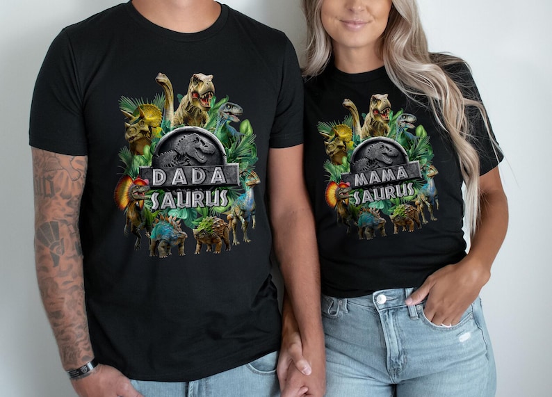 Peut inclure: T-shirts noirs avec des graphiques de dinosaures. L'un des t-shirts dit "DADA SAURUS" et l'autre "MAMA SAURUS". Les t-shirts pr&eacute;sentent un design inspir&eacute; de Jurassic Park avec diverses illustrations de dinosaures et du feuillage vert.