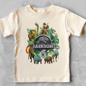 Peut inclure: T-shirt cr&egrave;me &agrave; manches courtes avec un motif de dinosaures color&eacute;s. Le design comprend le texte "JACKSON" et le chiffre "6" dans un logo sur le th&egrave;me de Jurassic Park, entour&eacute; de diverses illustrations de dinosaures.