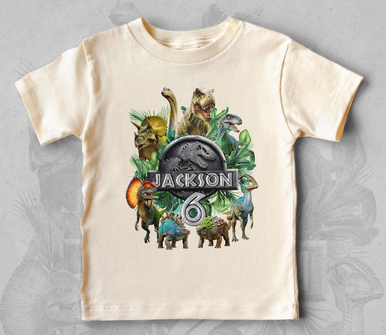 Peut inclure: T-shirt cr&egrave;me &agrave; manches courtes avec un motif de dinosaures color&eacute;s. Le design comprend le texte "JACKSON" et le chiffre "6" dans un logo sur le th&egrave;me de Jurassic Park, entour&eacute; de diverses illustrations de dinosaures.