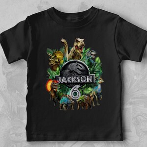 Peut inclure: T-shirt noir avec un motif de dinosaures et le texte "JACKSON 6". Le motif comprend divers dinosaures et le logo de Jurassic Park. Le t-shirt est fait d'une mati&egrave;re douce.