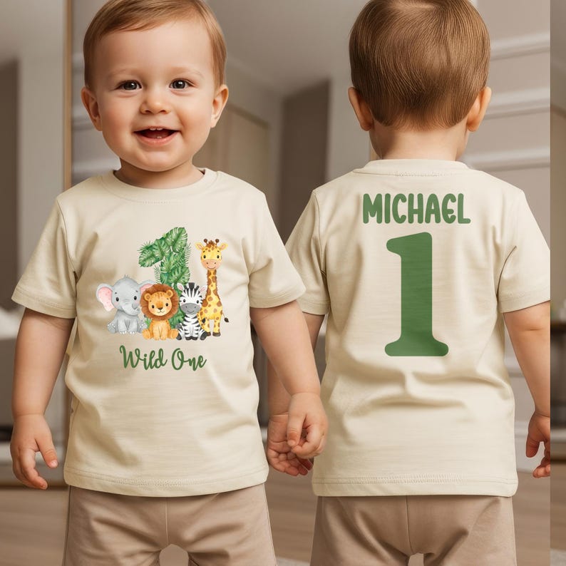 Pu&ograve; includere: T-shirt beige con grafica di animali della savana colorati e la scritta "Wild One". Il retro della maglietta riporta il nome "Michael" e il numero "1" in verde. La maglietta &egrave; indossata da un bambino piccolo.