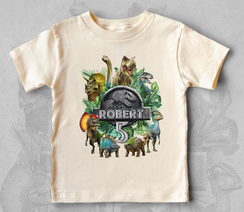 Peut inclure: T-shirt cr&egrave;me &agrave; manches courtes avec un motif de dinosaures color&eacute;s. Le design comprend divers dinosaures, le nom "ROBERT" et le chiffre "5" dans une police stylis&eacute;e, sur fond de feuillage vert.