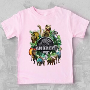Peut inclure: T-shirt rose clair avec un motif de dinosaures color&eacute;s, des feuilles vertes et le texte "ANDREW 7". Le design comprend un logo Jurassic Park. C'est un t-shirt &agrave; col rond et &agrave; manches courtes.
