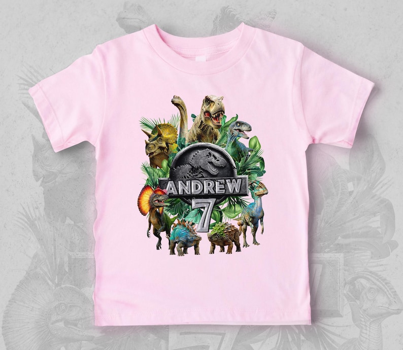 Peut inclure: T-shirt rose clair avec un motif de dinosaures color&eacute;s, des feuilles vertes et le texte "ANDREW 7". Le design comprend un logo Jurassic Park. C'est un t-shirt &agrave; col rond et &agrave; manches courtes.