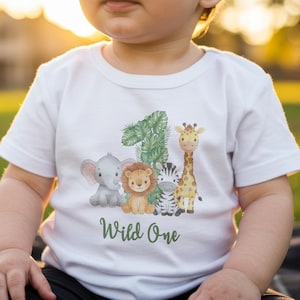 Pu&ograve; includere: T-shirt bianca a maniche corte con grafica a tema giungla. Il design include un elefante, un leone, una zebra e una giraffa, insieme al numero "1" e alle parole "Wild One" in verde. La maglietta &egrave; per bambini.