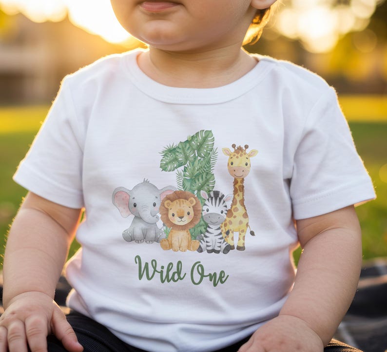 Pu&ograve; includere: T-shirt bianca a maniche corte con grafica a tema giungla. Il design include un elefante, un leone, una zebra e una giraffa, insieme al numero "1" e alle parole "Wild One" in verde. La maglietta &egrave; per bambini.