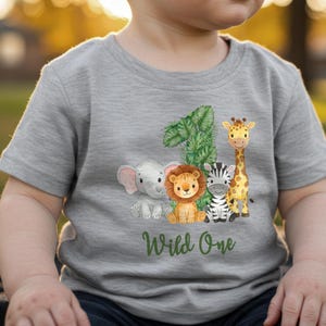 Pu&ograve; includere: Una maglietta grigia con un'illustrazione colorata del numero 1, una giraffa, una zebra, un leone e un elefante. Le parole "Wild One" sono scritte in verde sotto.