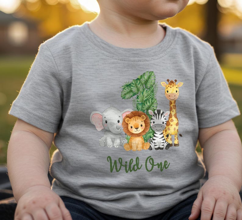 Pu&ograve; includere: Una maglietta grigia con un'illustrazione colorata del numero 1, una giraffa, una zebra, un leone e un elefante. Le parole "Wild One" sono scritte in verde sotto.