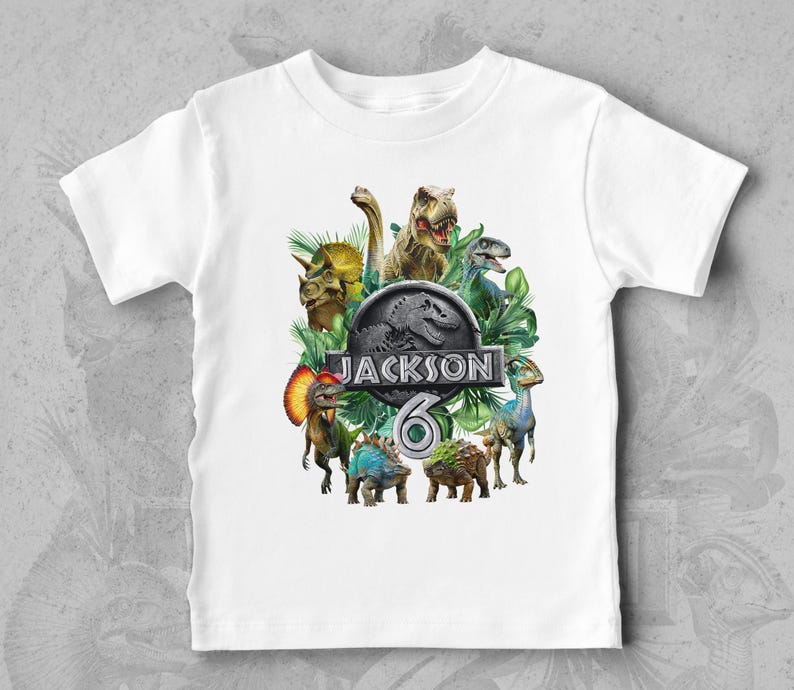 Peut inclure: T-shirt blanc avec un graphisme color&eacute; de dinosaures, de feuilles vertes et le texte "JACKSON 6". Le motif comprend un T-Rex, un Tric&eacute;ratops et d'autres cr&eacute;atures pr&eacute;historiques. Un design amusant et ludique pour enfants.