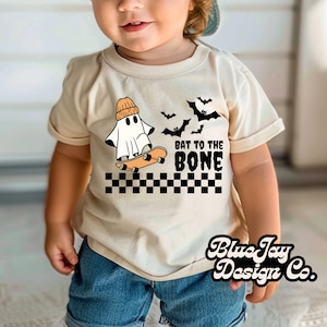 May include: A toddler wearing a tan t-shirt with a ghost skateboarding and the text "Bat to the Bone". The ghost is wearing a beanie and there are bats flying in the background. The shirt has a black and white checkered pattern at the bottom.
