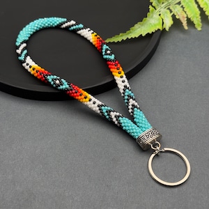 May include: A beaded key chain with a turquoise, white, black, red, and yellow geometric pattern. The key chain has a silver metal ring and a silver metal clasp.