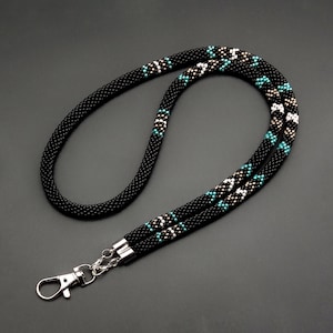 May include: Black beaded lanyard with a pattern of white and turquoise beads. The lanyard has a silver-colored clasp and is designed to hold an ID badge or keys. The background is a dark gray.