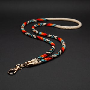 May include: A beaded lanyard featuring a woven pattern of white, black, orange, and turquoise beads. The cord is constructed from small, uniform beads and terminates in a gold-toned metal lobster clasp attached to a matching gold-toned metal end cap.