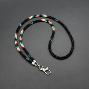 May include: Black beaded lanyard with a silver-colored clasp. The lanyard features a pattern of turquoise, brown, and white beads. The lanyard is designed to be worn around the neck.