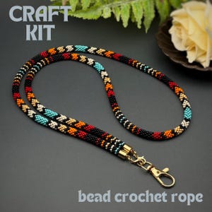 May include: A beaded crochet rope against a black backdrop. The rope features a repeating pattern of black, red, orange, turquoise, and white beads. It has gold-coloured metal clasps and the text "CRAFT KIT" and "bead crochet rope".