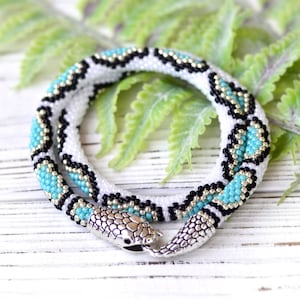 Peut inclure: Un collier de perles blanches avec des accents noirs et turquoise. Le collier est en forme de serpent et a un fermoir en forme de tête de serpent argenté.