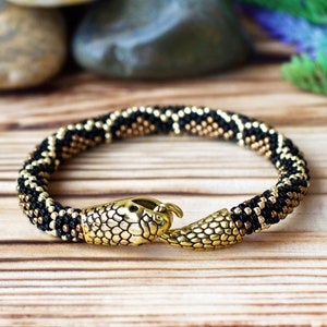 May include: A black and gold beaded bracelet with a snake head clasp. The bracelet is made of small, round beads and has a textured, snake-like pattern.