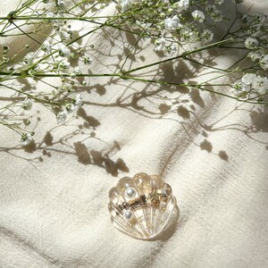 May include: A gold and white seashell-shaped hair clip with two pearl accents. The clip is on a white fabric background with white flowers and their shadows.