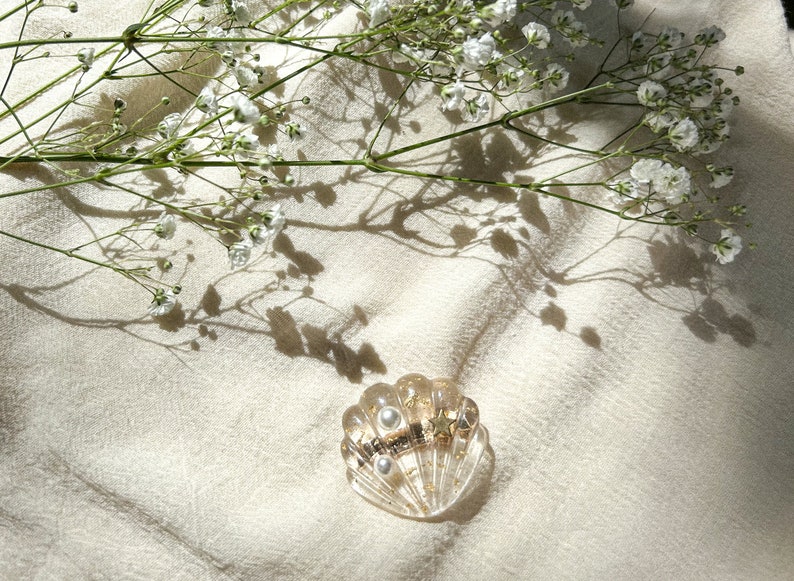 May include: A gold and white seashell-shaped hair clip with two pearl accents. The clip is on a white fabric background with white flowers and their shadows.